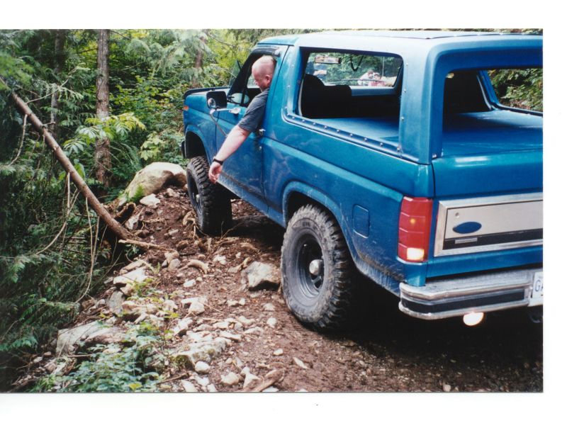 Blue 1980s Ford Bronco off-roading on a rocky, wooded trail.