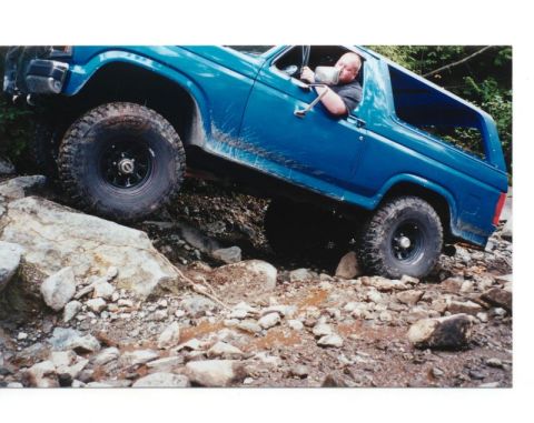 1985 Ford Bronco - A blue Ford Bronco is navigating a rocky, muddy trail with a person driving. The vehicle is angled, with its front tire on a large rock.