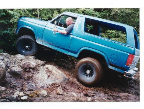 1985 Ford Bronco - A blue Ford Bronco is navigating a rocky, muddy off-road trail with a driver visible inside. The vehicle is angled steeply uphill, with its…