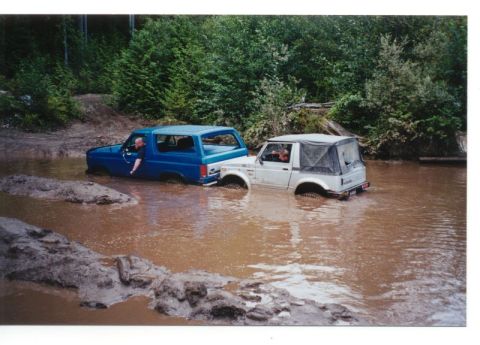 1985 Ford Bronco - A blue 1985 Ford Bronco and a white Suzuki Samurai are partially submerged in a muddy body of water, surrounded by trees.