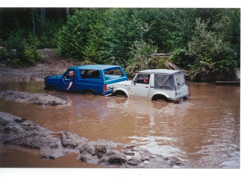 A blue Ford Bronco and a white Suzuki Samurai driving through a muddy water obstacle.