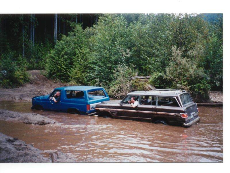 Blue Ford Bronco and brown Jeep Grand Wagoneer off-roading in mud and water.