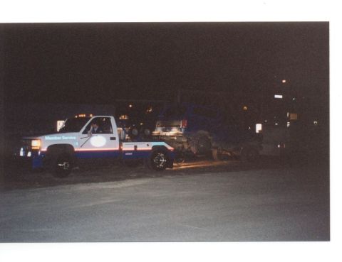 1985 Ford Bronco - A white and blue tow truck is shown at night, towing a blue SUV. The tow truck has "Member Service" written on the side.