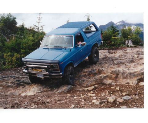 1985 Ford Bronco - A blue 1985 Ford Bronco is navigating a rocky, off-road trail with steep inclines and dense green foliage on either side. The vehicle has a…