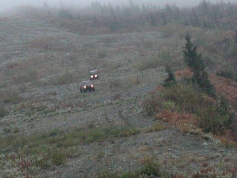 1985 Ford Bronco - Two SUVs are driving down a rocky, sparsely vegetated slope on a foggy day. The lead vehicle's headlights are on, illuminating the terrain…