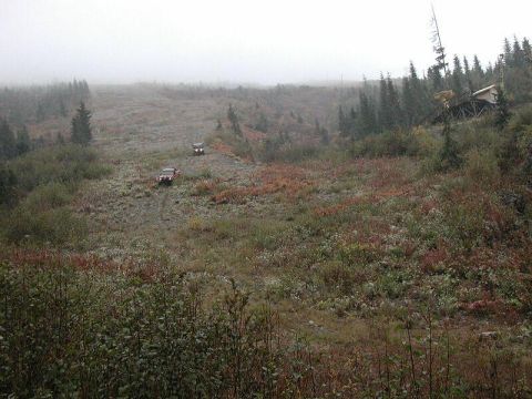 1985 Ford Bronco - Two off-road vehicles are driving up a muddy, rocky incline on a foggy day. A wooden structure is visible on the hillside to the right.