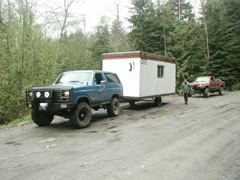1985 Ford Bronco - A blue Ford Bronco tows a white trailer with a small window down a dirt road, flanked by trees. A red pickup truck is parked behind the tra…