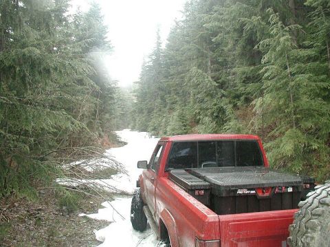 1985 Ford Bronco - The rear-left quarter view of a red pickup truck is shown from a low angle, driving on a snowy, wooded trail. The truck bed is equipped wit…