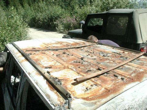 1985 Ford Bronco - The heavily rusted and burnt roof of a white SUV is shown in the foreground, with a dark green Jeep visible behind it on a dirt road. A per…