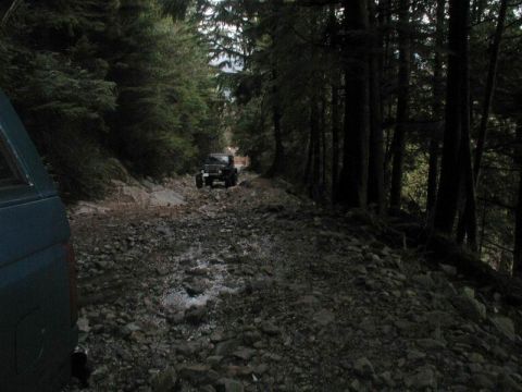 1985 Ford Bronco - A dark-colored Jeep is driving on a rocky, wooded trail. The front of a dark blue SUV is visible in the foreground on the left.