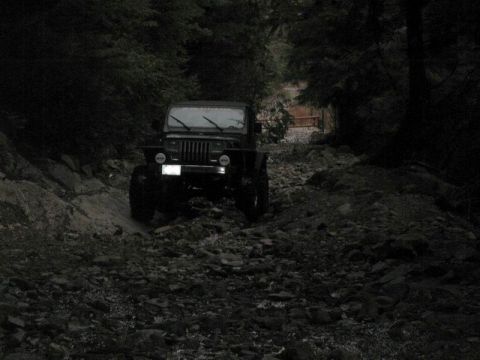 1985 Ford Bronco - A dark-colored Jeep Wrangler is navigating a rocky, wooded trail. The vehicle is facing forward, partially obscured by the low light and de…
