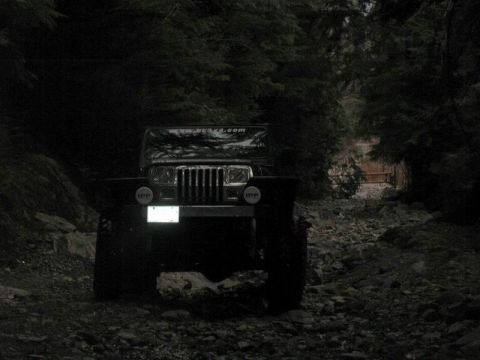 1985 Ford Bronco - A dark, front-facing view of a Jeep Wrangler driving on a rocky, wooded trail at dusk. The Jeep has IPF lights mounted on the front bumper…