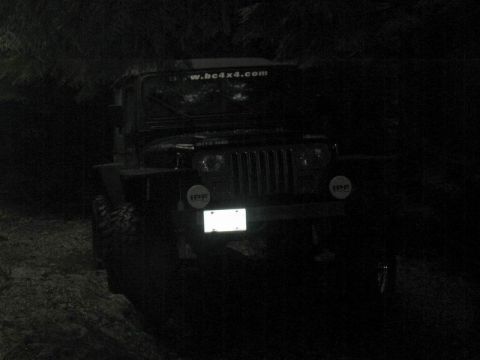 1985 Ford Bronco - A dark, nighttime photo shows the front of a black Jeep Wrangler with off-road lights. The vehicle is parked on a dirt path with trees in t…