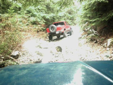 1985 Ford Bronco - A red pickup truck with a spare tire mounted on the back navigates a rocky, wooded trail. The front of another vehicle, possibly a boat or…