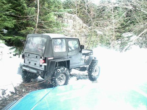 1985 Ford Bronco - A dark-colored Jeep Wrangler with a soft top is parked in a snowy wooded area. A "For Sale" sign is visible in the rear passenger window.