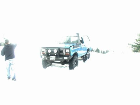 1985 Ford Bronco - A blue Ford Bronco is parked in deep snow, with a person standing to the left of the vehicle. The Bronco has large tires and a snorkel inta…