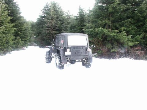 1985 Ford Bronco - A modified Jeep Wrangler with large off-road tires drives down a snow-covered path lined with tall evergreen trees.