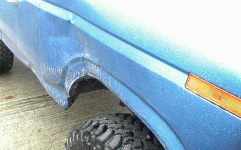 1985 Ford Bronco - Close-up side view of the front fender and wheel well of a blue SUV, possibly a Ford Bronco, with water droplets on the paint. The vehicle…