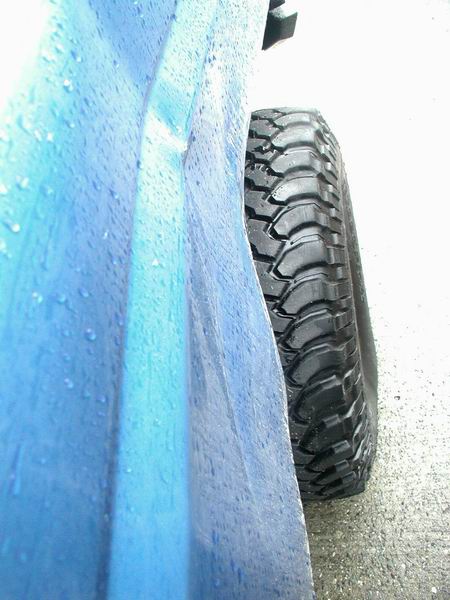 1985 Ford Bronco - A close-up side view of a wet, blue vehicle body panel next to a knobby off-road tire. The tire tread is aggressive and designed for off-ro…