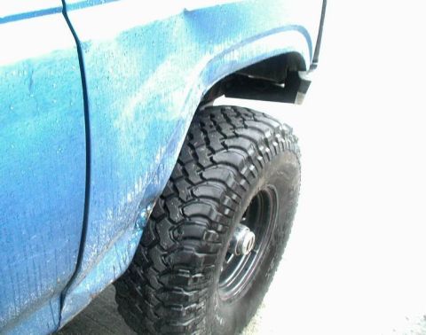 1985 Ford Bronco - A close-up, low-angle shot shows the rear fender and off-road tire of a blue Ford Bronco. The vehicle appears to be wet, with water droplet…