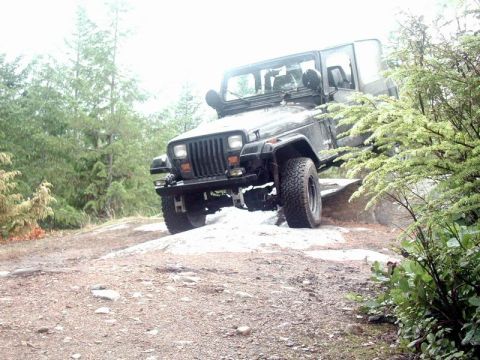 1985 Ford Bronco - A dark-colored Jeep Wrangler is navigating a rocky, off-road trail with dense trees in the background. The front of the Jeep is angled towa…