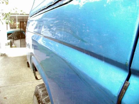 1985 Ford Bronco - A close-up, side view of the rear fender and door of a blue Ford Bronco. The vehicle appears to be parked outdoors on a concrete surface.