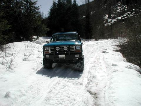 1985 Ford Bronco - A teal 1985 Ford Bronco is parked on a snow-covered dirt road surrounded by trees. The vehicle has a black aftermarket bumper with IPF ligh…