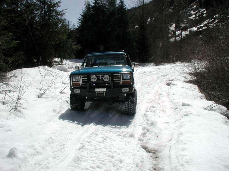 Teal 1985 Ford Bronco on a snowy road with aftermarket bumper and lights.