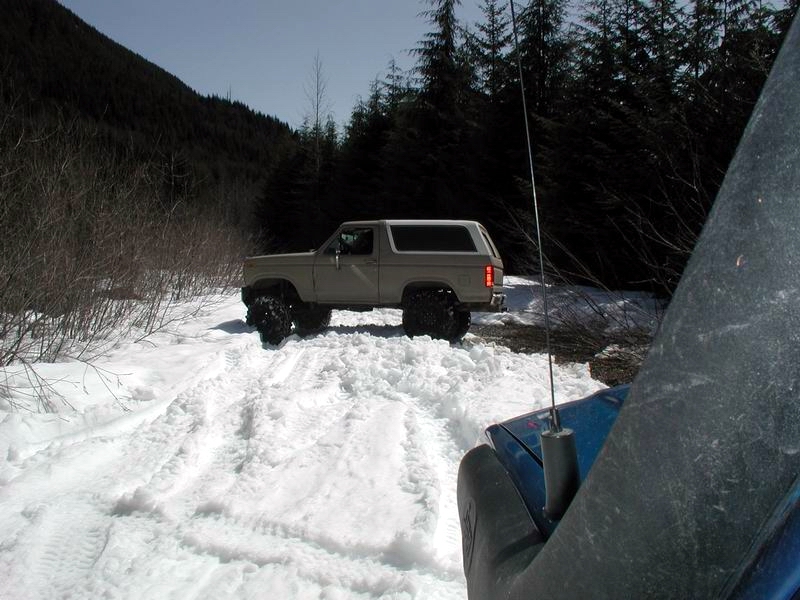Tan 1985 Ford Bronco with large tires driving on a snowy trail.