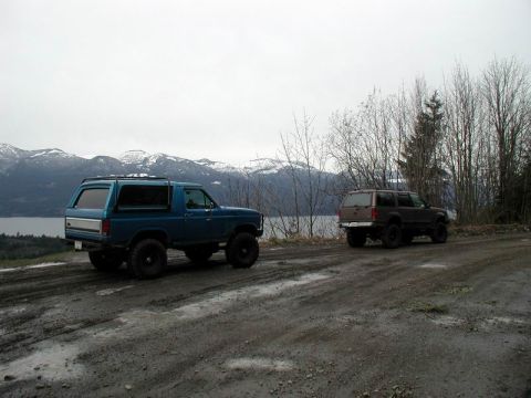 1985 Ford Bronco - A lifted blue 1980s Ford Bronco is parked on a muddy dirt road, with a dark SUV parked behind it. Snow-capped mountains and a body of water…