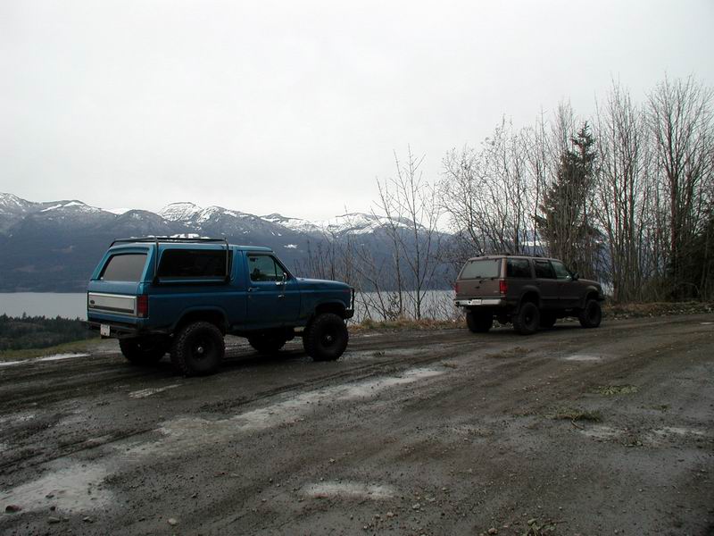 Lifted blue 1980s Ford Bronco and dark SUV on a muddy road with mountains in background.