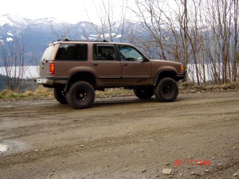 1985 Ford Bronco - A lifted brown Ford Explorer is parked on a dirt road with a lake and snow-capped mountains in the background.