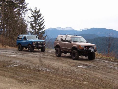 1985 Ford Bronco - Two modified SUVs are parked on a dirt road with mountains in the background. The blue SUV on the left is a Ford Bronco, and the brown SUV…