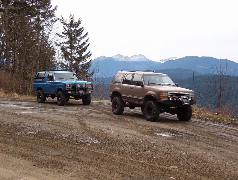 Blue Ford Bronco and brown Ford Explorer modified for off-roading on a dirt road with mountains.