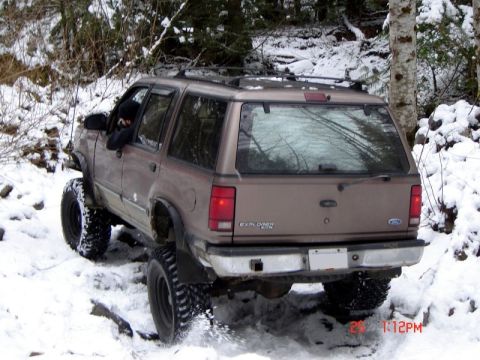 1985 Ford Bronco - A brown Ford Explorer is driving off-road through snow-covered terrain with large tires. The vehicle is angled to the left, showing its rea…