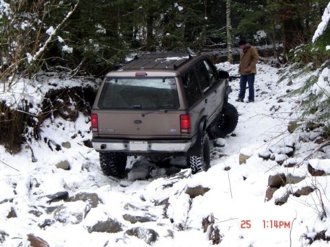 1985 Ford Bronco - A tan Ford Explorer is navigating a rocky, snow-covered trail in a wooded area. A person stands nearby watching the vehicle.
