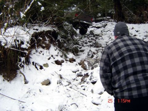 1985 Ford Bronco - A person wearing a plaid jacket and beanie stands in the foreground, looking towards a rocky, snow-covered trail where a brown Ford Bronco…