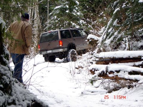 1985 Ford Bronco - A brown SUV is driving on a snow-covered trail in a wooded area. A person wearing a tan jacket and blue jeans stands to the left, looking t…