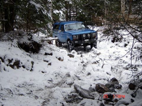 1985 Ford Bronco - A blue 1985 Ford Bronco navigates a rocky, snow-covered trail in a wooded area. The vehicle is equipped with off-road tires and auxiliary l…