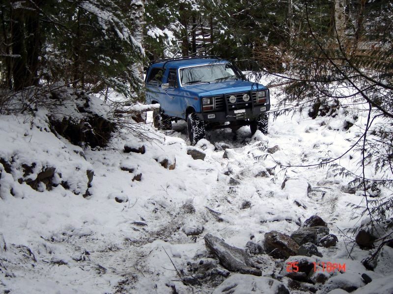 Blue 1985 Ford Bronco off-roading on a snowy, rocky trail.
