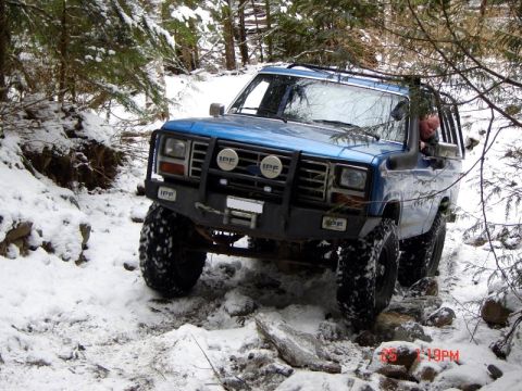1985 Ford Bronco - A blue Ford Bronco navigates a snowy, rocky trail surrounded by trees. The vehicle has a prominent brush guard with IPF lights and large of…