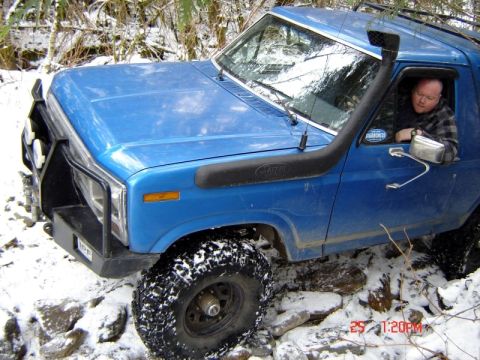 1985 Ford Bronco - A blue, 1980s Ford Bronco is navigating a snowy, rocky trail with a snorkel installed. The driver is visible through the window, looking to…