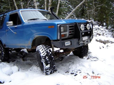 1985 Ford Bronco - A blue Ford Bronco is shown off-road in a snowy forest. The vehicle has a custom front bumper with IPF lights and a winch.