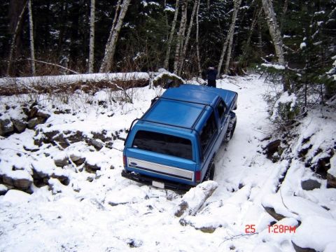 1985 Ford Bronco - A blue Ford Bronco is navigating a snowy, wooded trail with a person standing in the distance. The vehicle is angled away from the camera,…