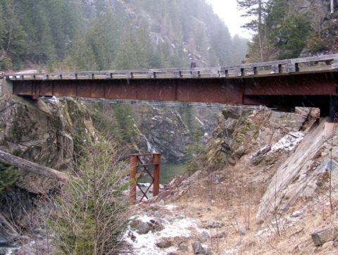 1985 Ford Bronco - A rusty metal bridge spans a rocky gorge with a river below. The scene is set in a wooded, mountainous area with snow flurries falling.