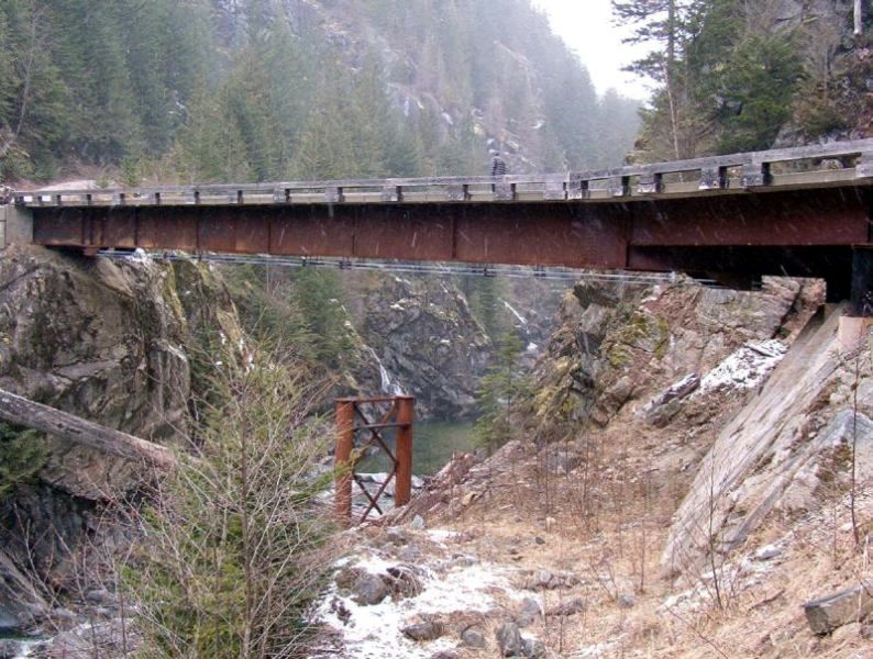 A rusty metal bridge over a rocky gorge in a snowy, wooded mountain landscape.