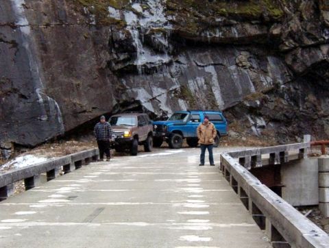 1985 Ford Bronco - Two men stand on a bridge next to a brown SUV and a blue SUV, with a rocky cliff face behind them.