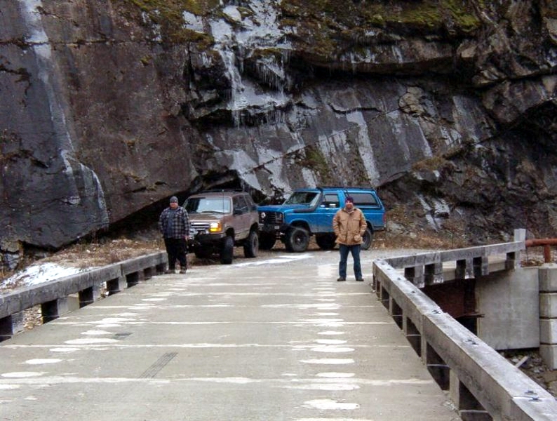 Two men and two SUVs on a bridge with a rocky cliff face.