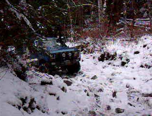 1985 Ford Bronco - A blue 1985 Ford Bronco is navigating a snowy, wooded trail. The vehicle is angled towards the viewer's left, with its front grille and hea…