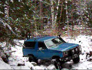 1985 Ford Bronco - A blue 1985 Ford Bronco is parked in a snowy forest with trees and snow-covered ground visible. The vehicle has a snorkel and a brush guard…